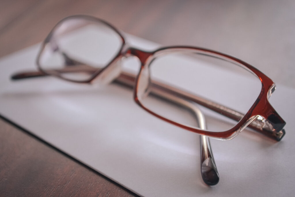 Image of reading glasses the colour red-brown. The glasses are resting on a dark brown wooden table on top of a white piece of paper (presumably an envelope)
